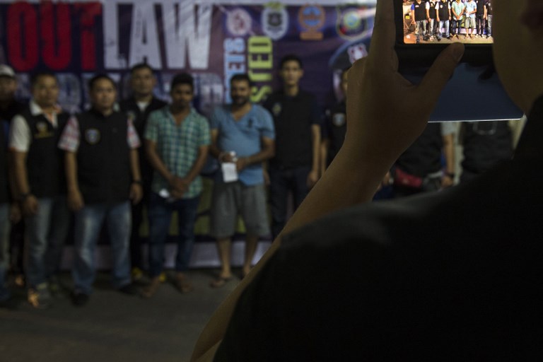 This picture taken on October 11, 2018 shows police and foreigners held for questioning at Bangkok’s Nana district posing before a police officer for a picture during a police operation called “X-Ray Outlaw Foreigner”. – Allegedly aimed at busting visa abusers and illegal migrants, a Thai police operation called “X-Ray Outlaw Foreigner” has raised questions about racial profiling and fears for asylum-seekers caught in its web. (Photo by Romeo GACAD / AFP)