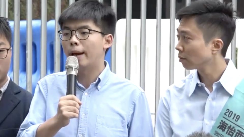 Joshua Wong (left) speaks to reporters hours after a returning officer barred him from running in the upcoming district council elections on Nov. 24. He urged voters to support the pro-democracy side’s ‘plan B candidate’ Kelvin Lam (right). Screengrab via YouTube/Apple Daily.